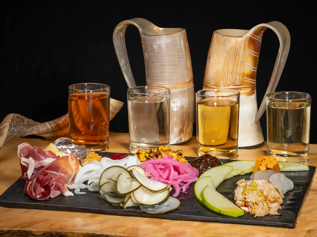 Black platter with charcuterie, cheese, pickles, drinks, and horn pitchers.