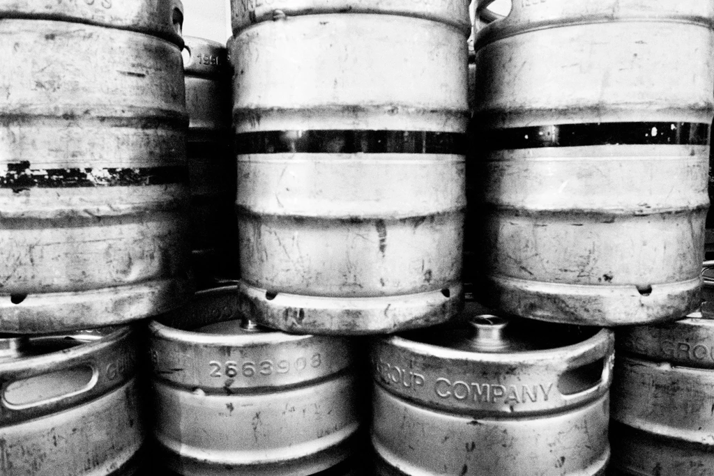 Dense stack of worn metal beer kegs in black and white.