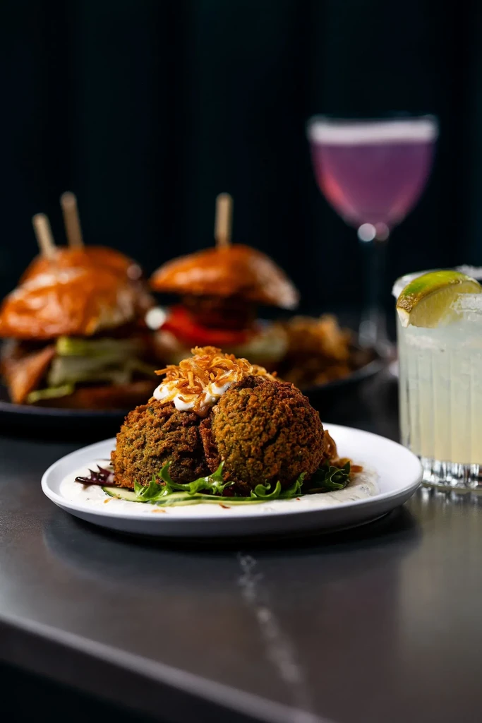 Falafel-like dish, cocktail, and blurred mini-burgers and drinks on table.