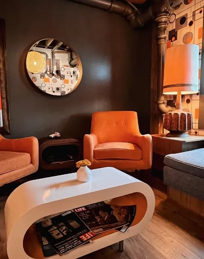 Mid-century modern lounge with orange armchairs, coffee table, and geometric wallpaper.