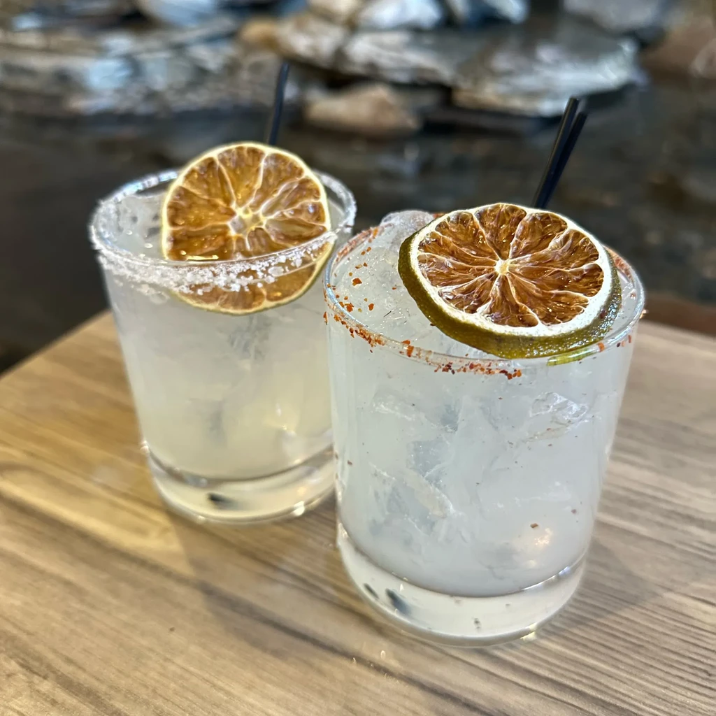 Two cocktails with ice, dried citrus, salted and spiced rims on wood.