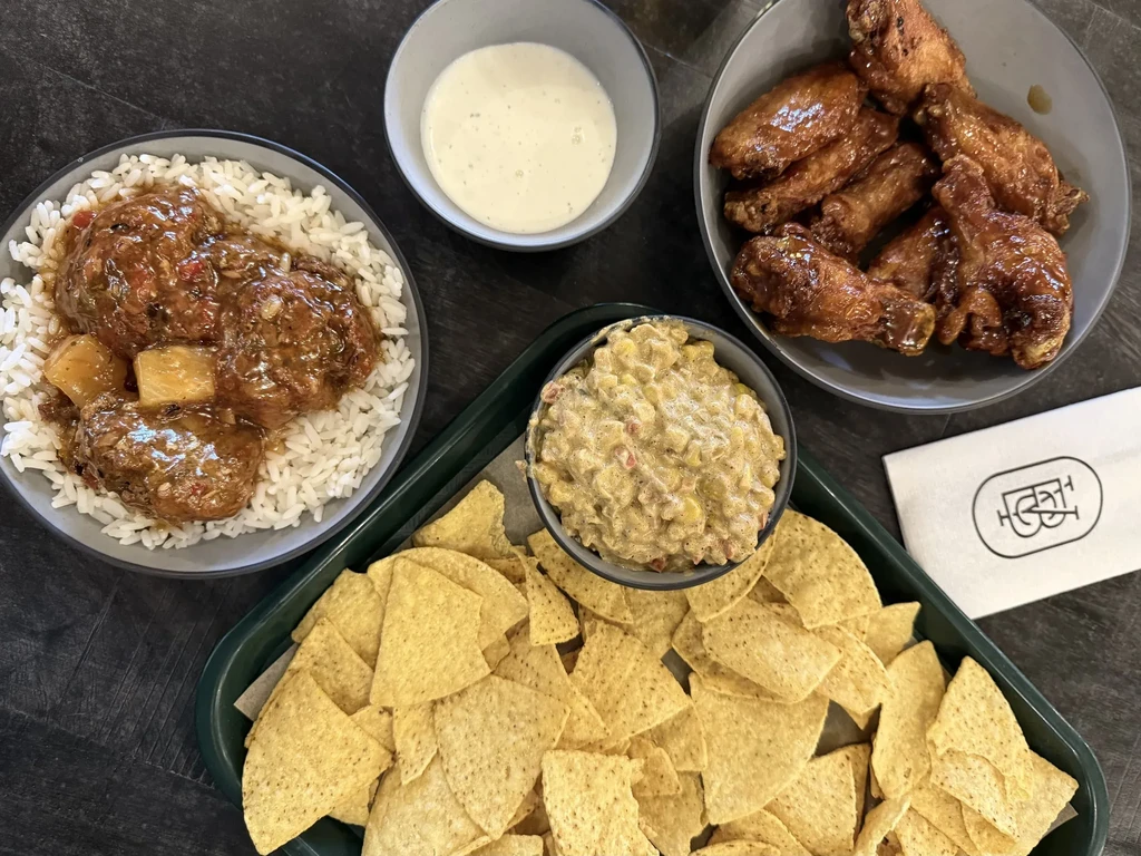 Assortment of sauced meatballs, chicken wings, chips, and dips on table.