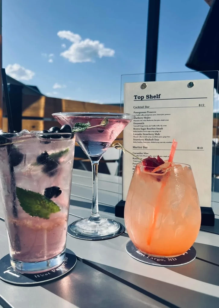 Three colorful cocktails on an outdoor table, with menu and blue sky.