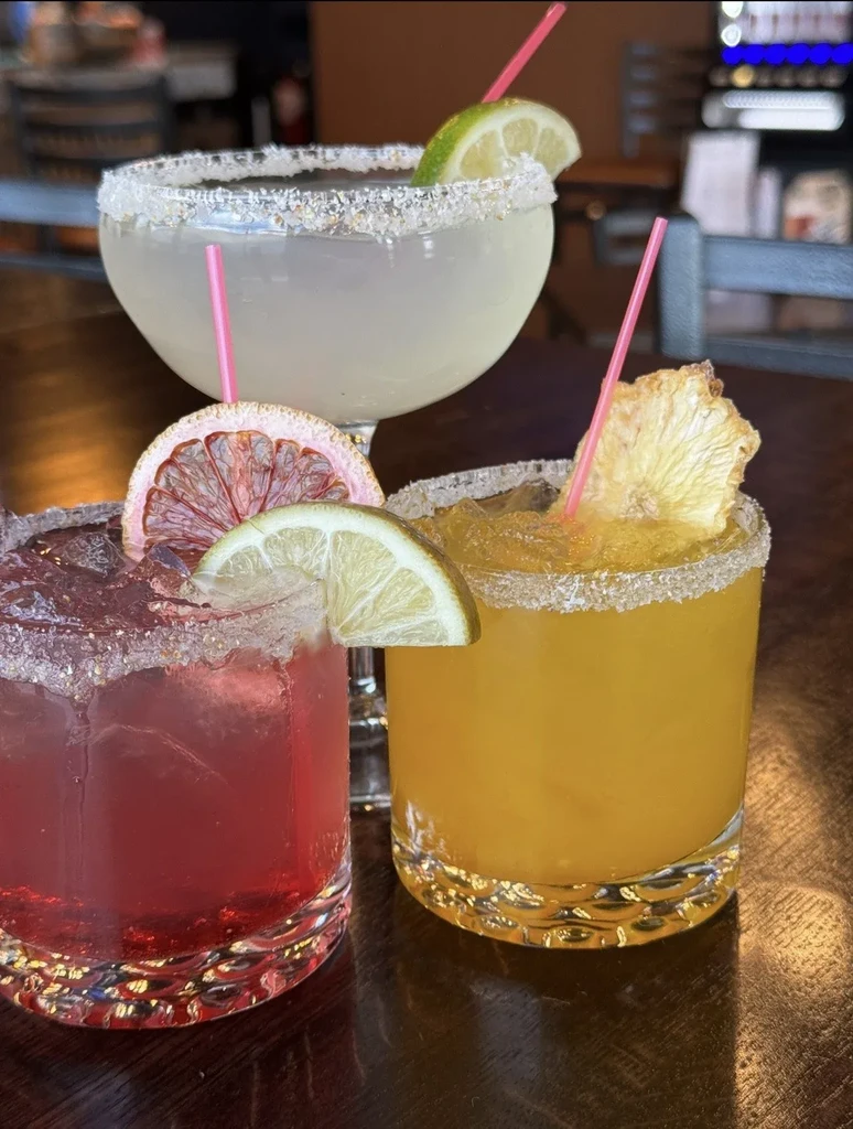 Three vibrant cocktails with garnishes and pink straws on a dark table.