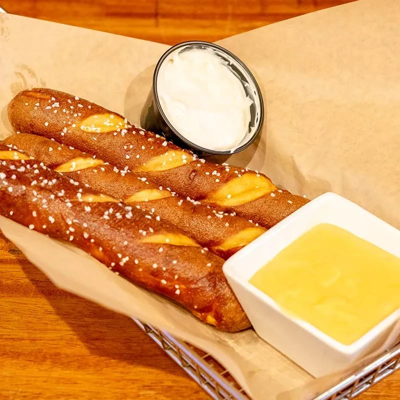 Three salted pretzel sticks in a basket, with white and yellow dips.