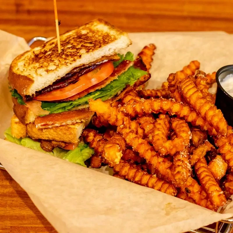 Stacked sandwich, curly fries, and dipping sauce served in a metal basket.