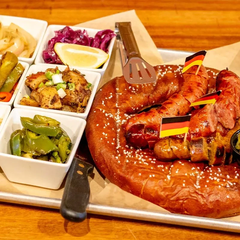 Pretzel and grilled sausages platter with German flags and side dishes.