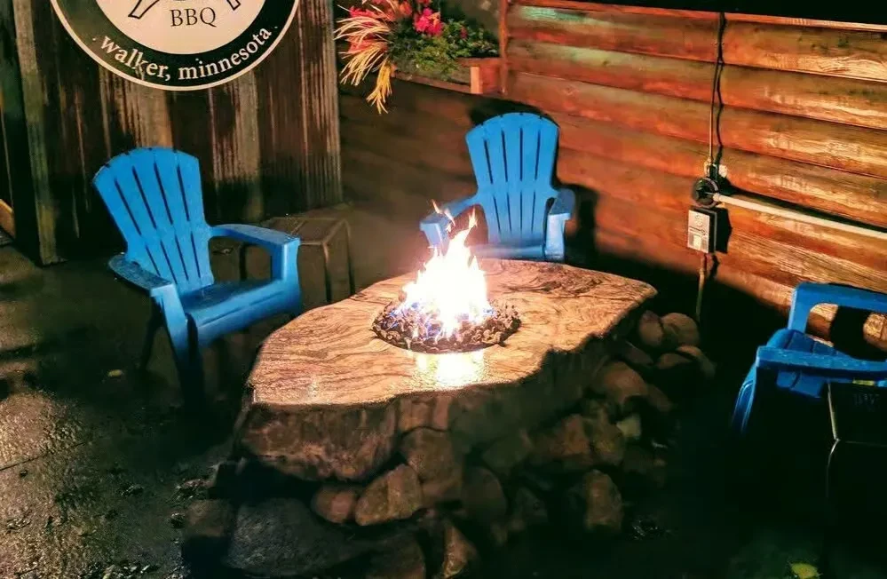 Outdoor stone fire pit table with flame and blue Adirondack chairs at night.