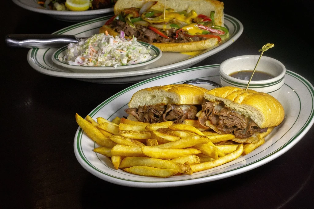 Sliced beef sandwich and cheesesteak with sides on a dark table.