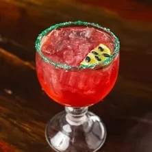 Red slushy drink in goblet with green rim and fruit garnish.