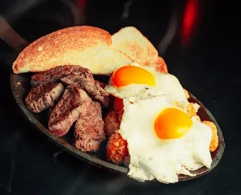 Close-up of seared steak, fried eggs, and toast on a dark skillet.
