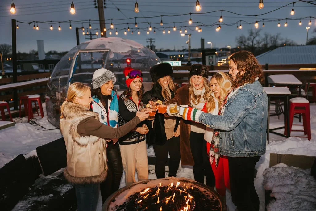 Seven people toast drinks around a snowy fire pit with an igloo.