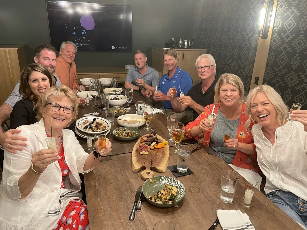 Nine smiling adults dine and drink around a rustic wooden table.