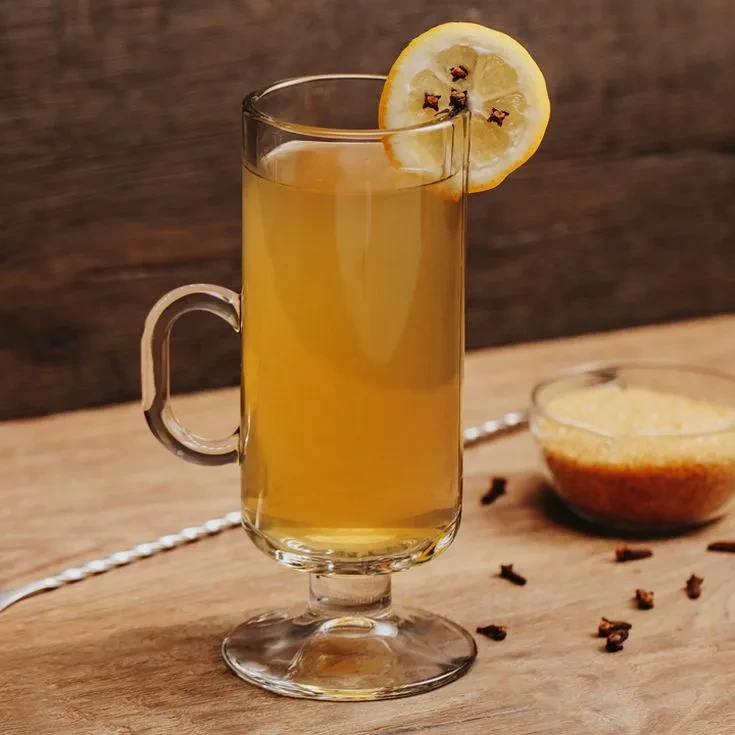 Golden-yellow drink with lemon and cloves in a clear mug on wood.