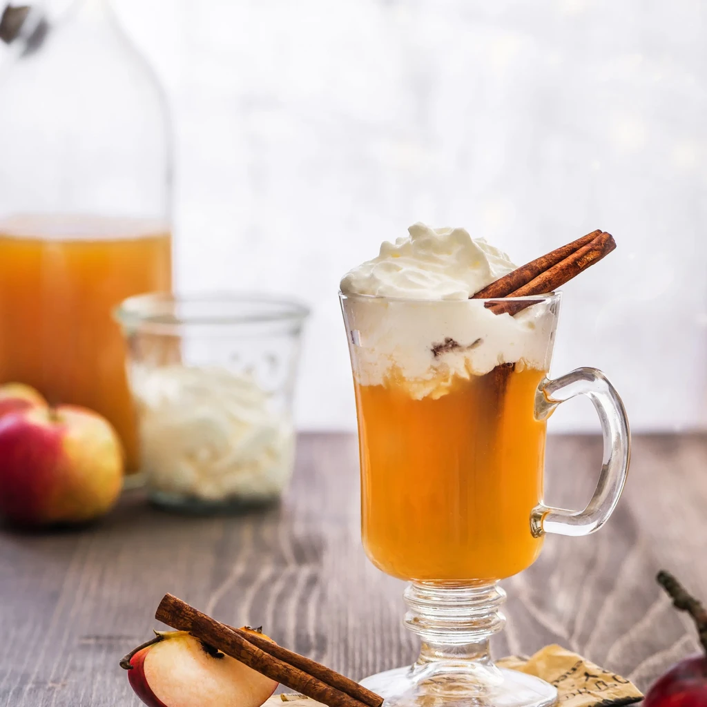 Mug of hot cider with whipped cream, cinnamon sticks, and apples.