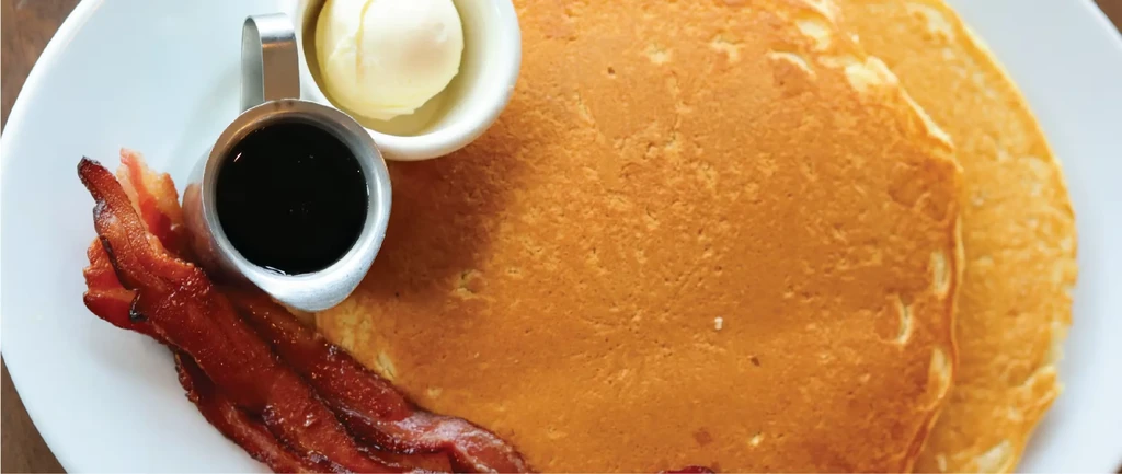 An overhead shot of pancakes, bacon, syrup, and butter on a plate.