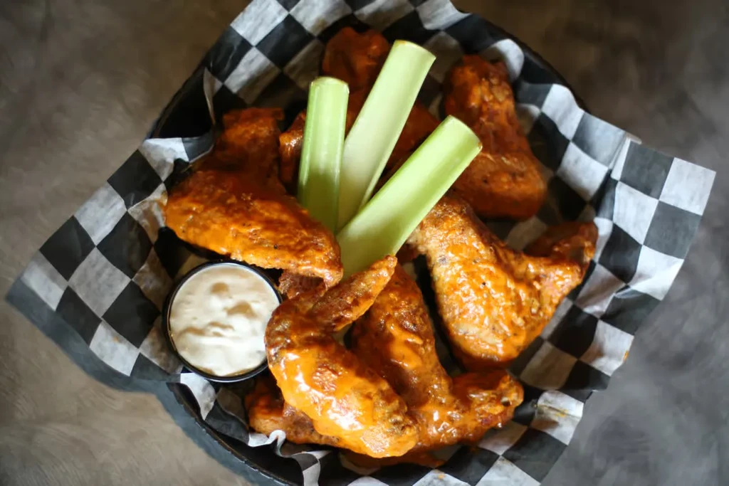 Glistening buffalo chicken wings, celery, and white dip in a checkered basket.