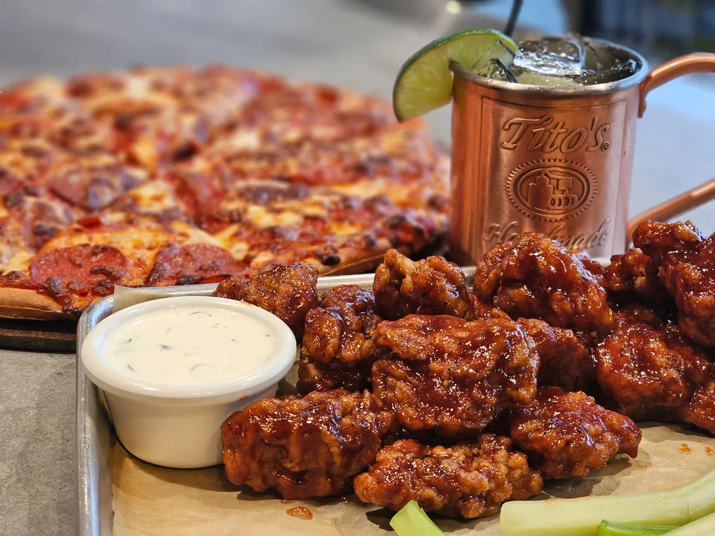 Saucy chicken wings, celery, blurred pizza, and Tito's Moscow Mule.
