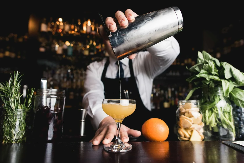 Bartender pours cocktail from silver shaker, surrounded by fresh ingredients.
