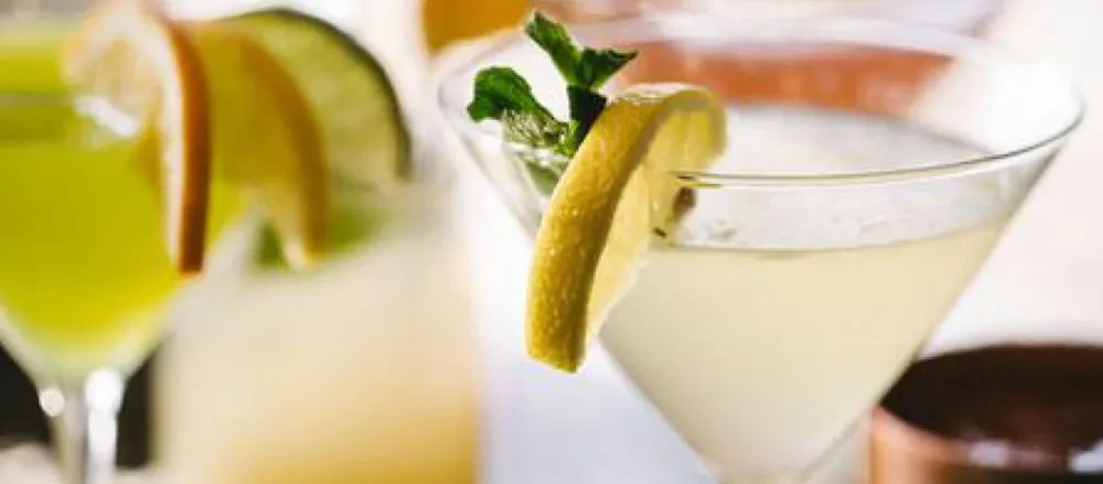 Pale yellow cocktail with lemon and mint, a green cocktail with citrus behind.