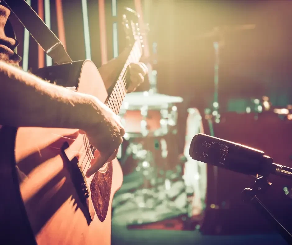 Hands strumming acoustic guitar on stage with drums and microphone visible.