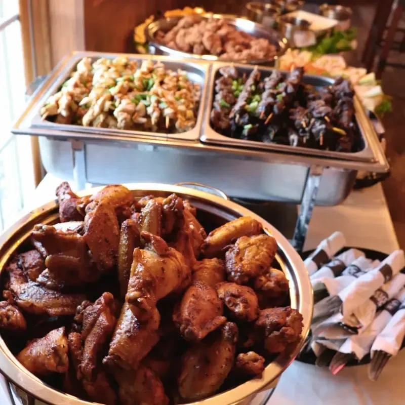 Buffet with chafing dishes of chicken wings and meat skewers.