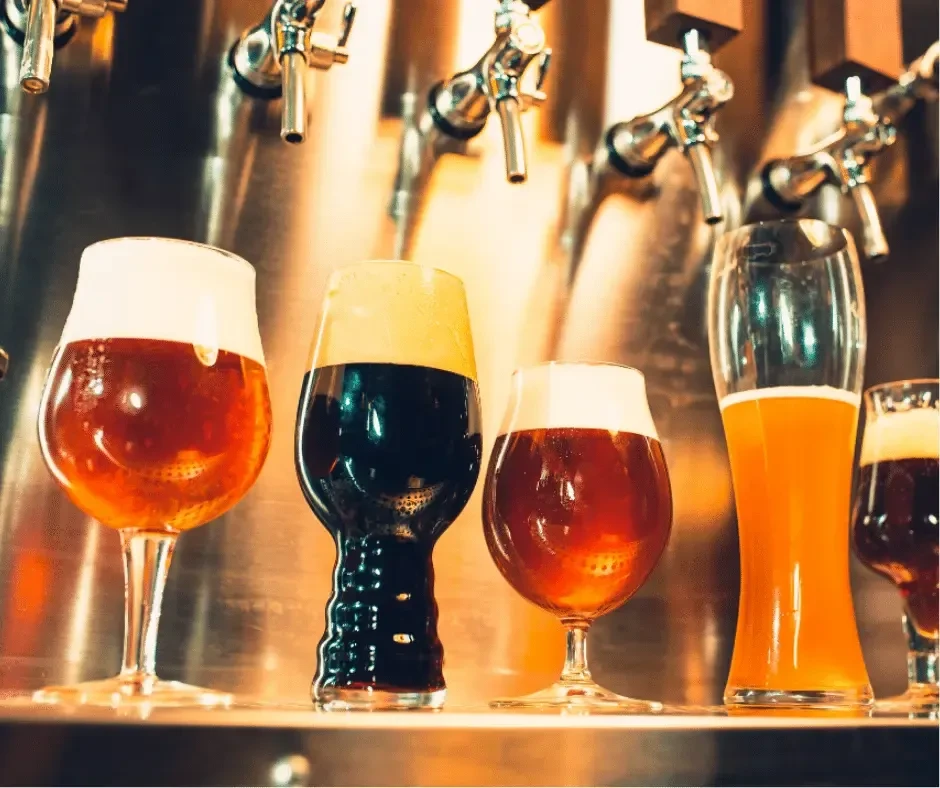 Five glasses of various beers on a bar, near metallic taps.
