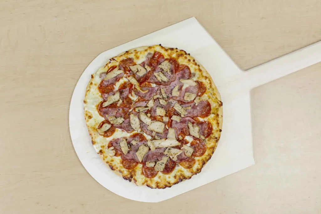 Round pizza with pepperoni, ham, chicken, herbs on wooden peel.