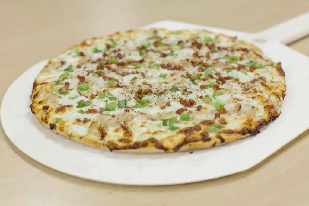 Freshly baked white sauce pizza with chicken, pepper, and bacon on a peel.