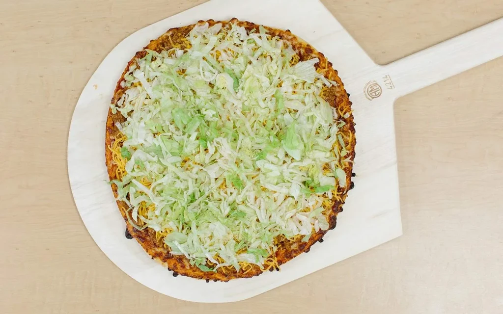 Round pizza topped with meat, cheese, and shredded lettuce on a wooden peel.
