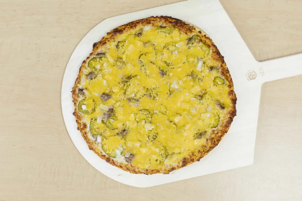 Round pizza with cheese, ground meat, pickles, and onions on a wooden peel.