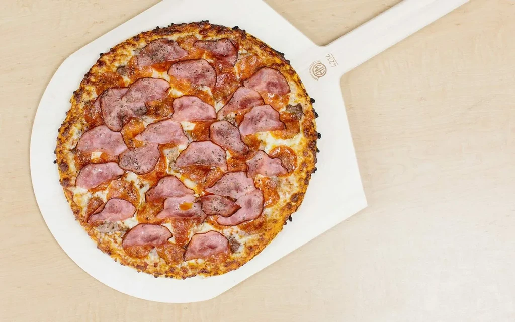 Top-down pizza with pepperoni, ham, and cheese on a white peel.