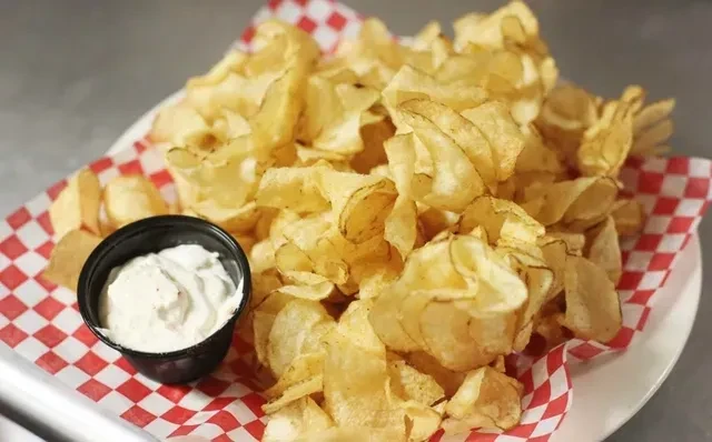 Plate of golden potato chips and creamy dip on checkered paper.