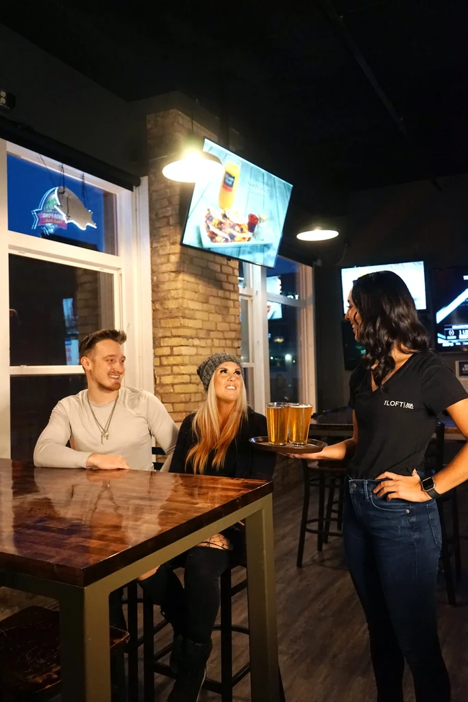 Server delivers two beers to a smiling couple in a lively restaurant.