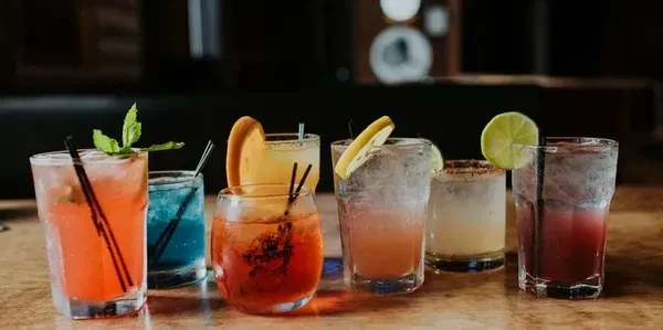 Seven colorful garnished cocktails in various glasses on a bar.