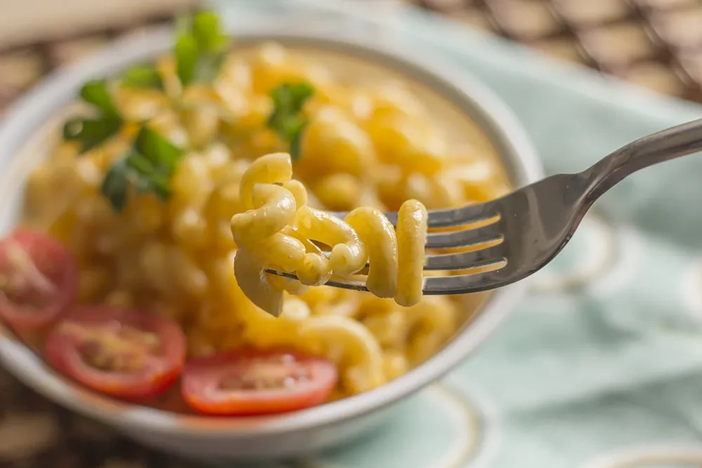 Fork lifting cheesy macaroni from a bowl, garnished with herbs and tomatoes.