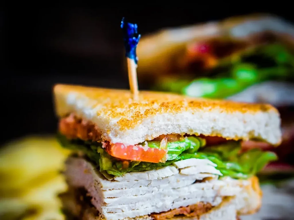 Stacked sandwich with meat, lettuce, tomato, toasted bread, and toothpick.