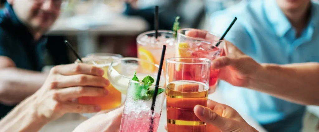 Hands clinking five colorful iced drinks with straws and garnishes.