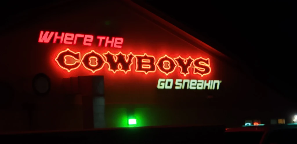 Ornate neon sign "WHERE THE COWBOYS GO SNEAKIN'" glowing in the dark.