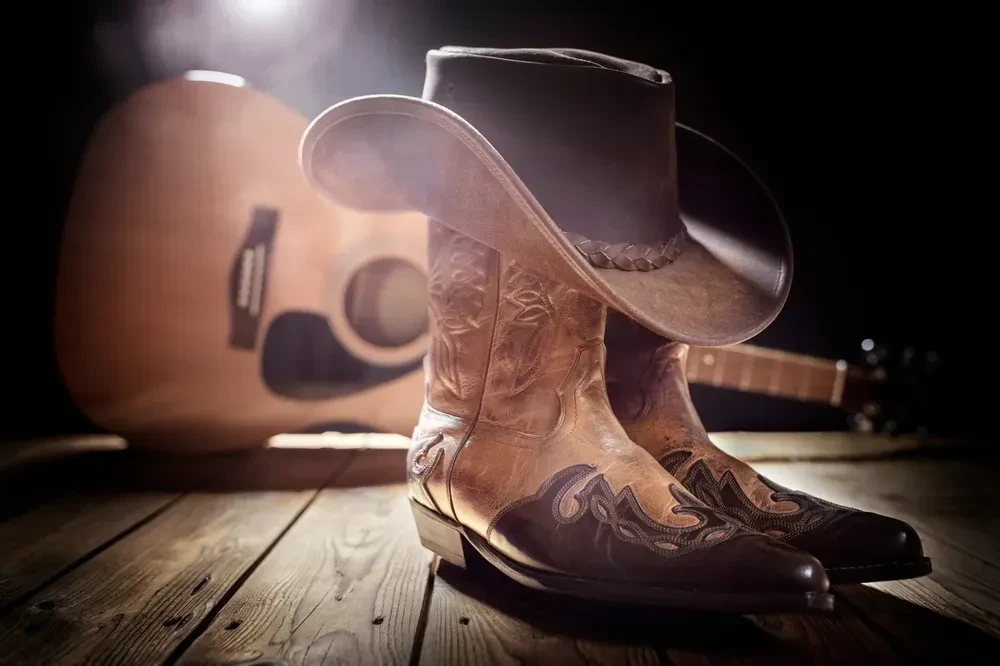 Spotlight on worn cowboy boots, hat, and guitar on a rustic floor.