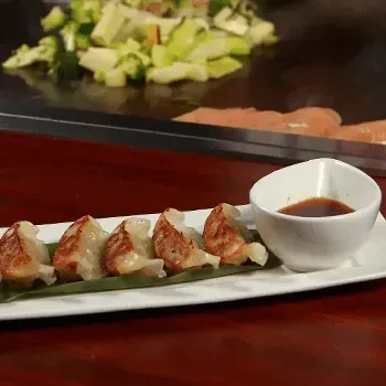 Five pan-fried gyoza dumplings with sauce, next to a cooking teppanyaki grill.