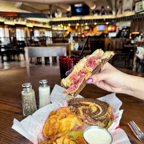 Hand holds Reuben sandwich, revealing layers, chips, and pickle.