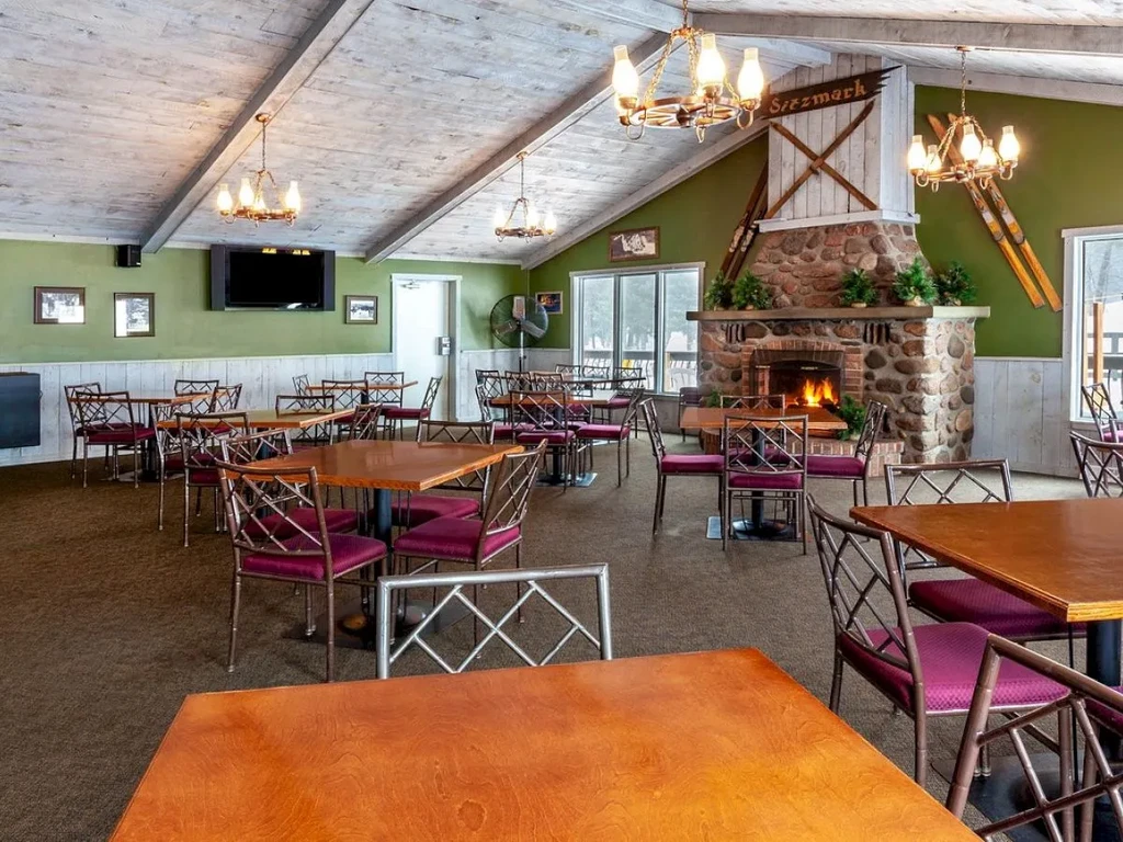 Rustic restaurant dining room with stone fireplace, wooden tables, and wagon-wheel chandeliers.