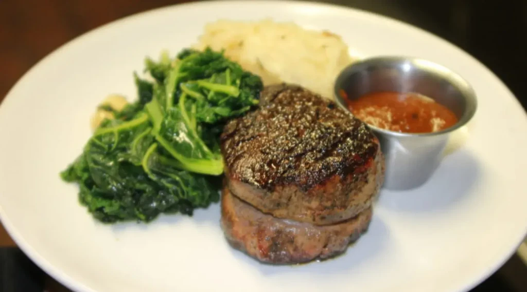 Plated medium-rare steak, green vegetables, mashed potatoes, and red sauce.
