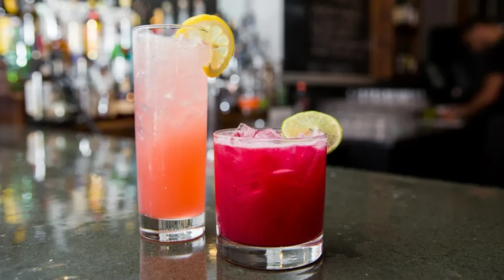 Pink and magenta cocktails with lemon and lime on a dark bar.