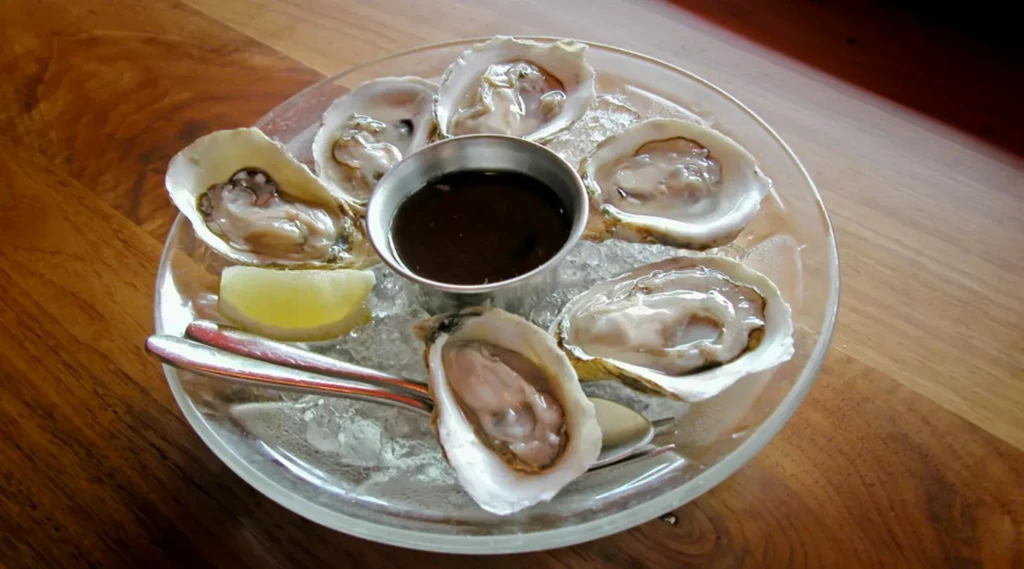 Six fresh oysters on ice with dipping sauce, lemon, and forks.