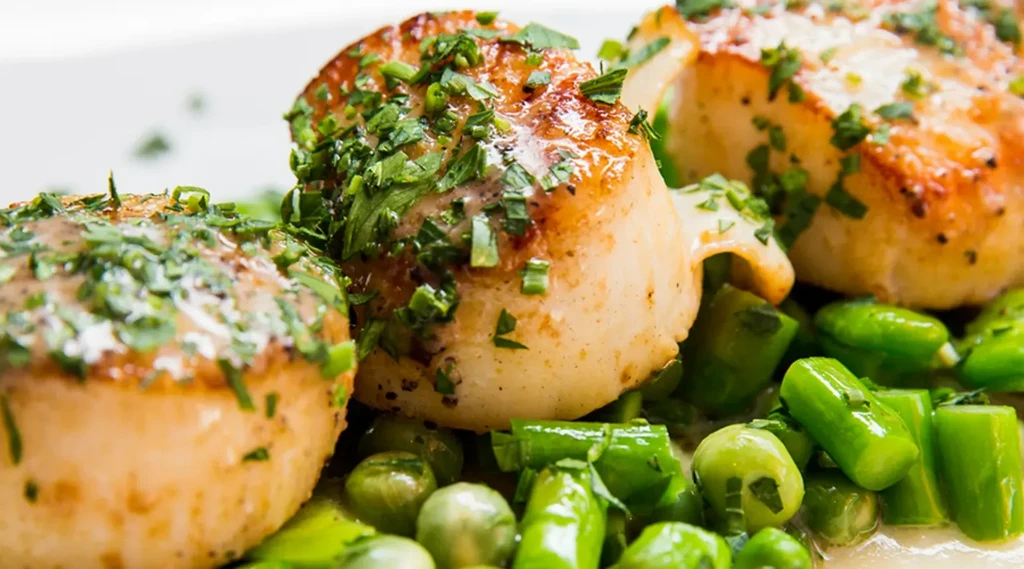 Three golden-brown seared scallops with herbs on green vegetables.