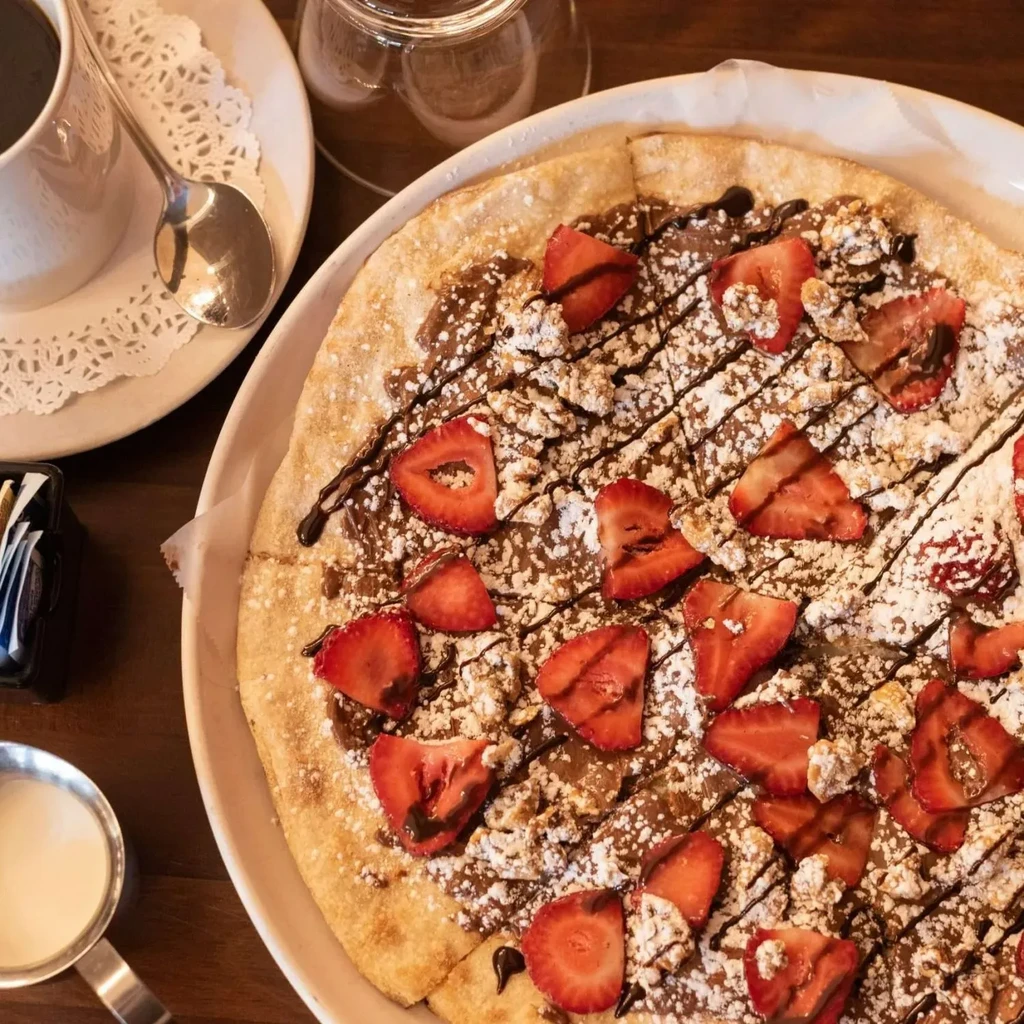 Overhead: Chocolate strawberry dessert pizza, coffee, creamer on wooden table.