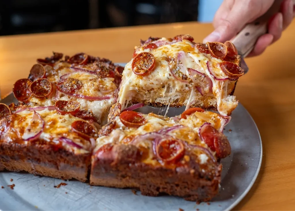 Hand lifts square deep-dish pizza slice, cheese stretching, pepperoni, onions.