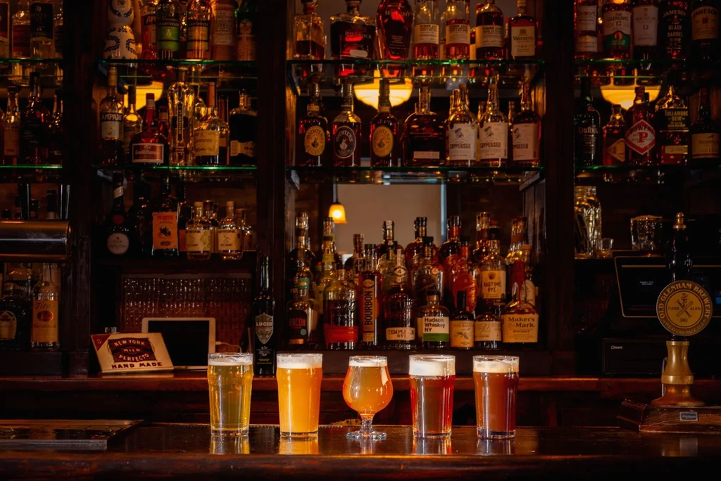 Five glasses of beer on a dark, well-stocked bar.